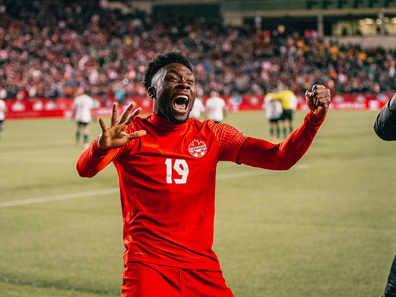 Soccer player in a red Canada jersey celebrates on the field with raised hands in a packed stadium
